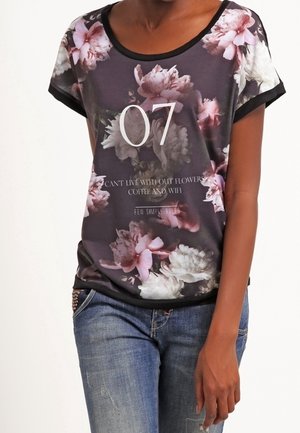 Kvinna klädd i en svart blommig t-shirt med "07" och text om blommor, kaffe och WiFi, i kombination med blå jeans.