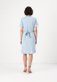 Femme aux cheveux courts et bouclés portant une robe bleu clair longueur genoux avec des manches courtes et une ceinture nouée à la taille, debout devant un fond uni.