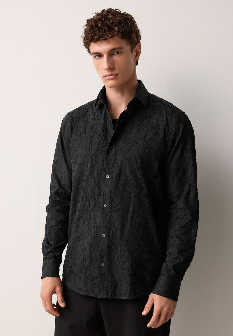 Schwarzes Langarmshirt mit strukturiertem Blumendesign, durchgeknöpft mit schwarzen Knöpfen und spitzem Kragen. Hergestellt aus atmungsaktivem Stoff.