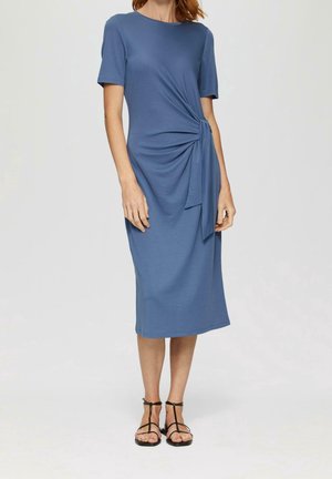 Woman wearing a knee-length blue dress with short sleeves and a side knot detail, paired with black strappy sandals.