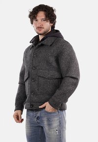 Giacca grigia in tessuto con bottoni frontali, grandi tasche e una felpa nera sotto. Presenta un colletto e un design angolato.