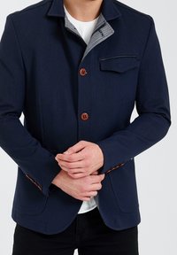 Marineblaue Jacke mit strukturiertem Design, aus Holzknöpfen, einer Fronttasche und einem grauen, gemusterten Kragenfutter.