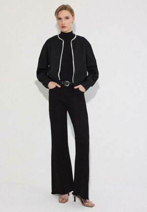 Femme portant un pantalon large noir taille haute, un haut noir à manches longues, une veste courte noire avec un bord blanc, et des sandales à talons beiges.