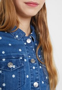 Close-up of woman wearing blue denim jacket with white polka dots and metal buttons, showcasing long light brown hair and partial smile.