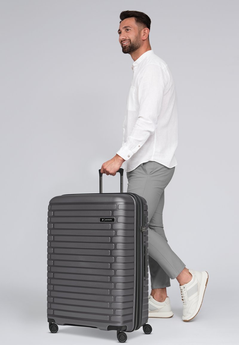 Valise rigide gris foncé avec des rainures horizontales, roues doubles et une poignée télescopique. Le nom de la marque est affiché de manière proéminente.