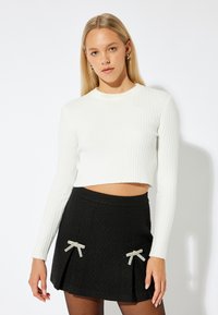 Weißes geripptes Crop-Top mit langen Ärmeln, kombiniert mit einem schwarzen strukturierten Rock, der mit zwei silbernen Schleifenakzenten versehen ist. Das Outfit ist figurbetont.