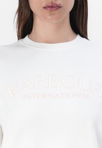 Fehér pulóver, kerek nyakkivágással és nagy, domború "BARBOUR INTERNATIONAL" felirattal, lágy barack színben, a mellkasán.