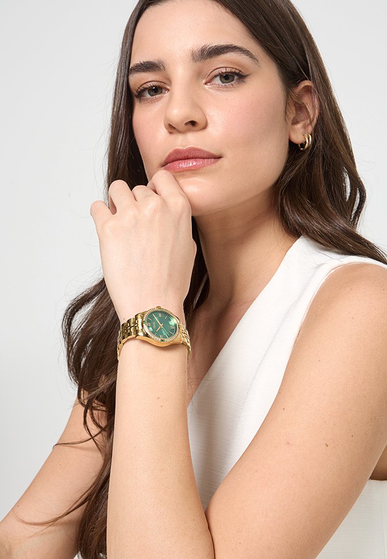 Montre en métal doré avec un cadran vert, présentant des index et des aiguilles dorées. La montre est équipée d'un bracelet à maillons et d'une lunette ronde.