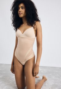 Naakt bodysuit met een gladde stof, V-hals ontwerp en verstelbare bandjes, met doorzichtige zijpanelen en een hoge snit aan de benen.
