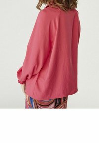 Löst sittande, långärmad skjorta i ett livligt rosa tyg med en texturerad yta. Har en volangfådd ärm och en böjd fåll, parat med mönstrade byxor.