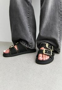 Sandales en daim noir avec des accents de double boucle dorée et des détails cloutés, présentant une semelle plate et un design ouvert au bout.