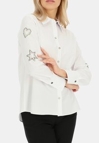 Biała koszula z długim rękawem, z ozdobami w kształcie serca i gwiazdy z rhinestones na łokciach oraz kontrastującymi czarnymi guzikami.