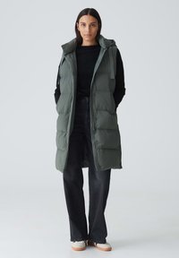 Langer, olivgrüner gepolsterter Weste mit Kapuze, mit Reißverschluss vorn und Seitentaschen, über einem schwarzen Pullover und schwarzen Jeans getragen.