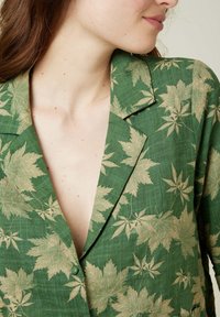 Chemise vert en tissu texturé avec un motif de feuilles dorées. Elle possède un col à revers et un décolleté en V profond.