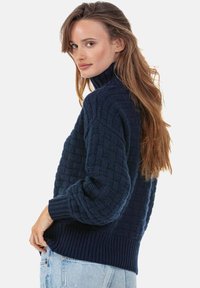 Pull tricoté bleu marine avec un col montant, présentant un motif texturé et des poignets côtelés. Conçu avec une coupe ample et des épaules tombantes.