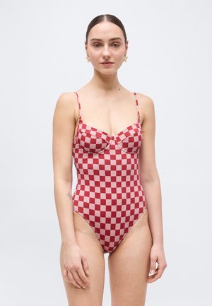 Γυναίκα με τα μαλλιά χτενισμένα προς τα πίσω, φορώντας σκουλαρίκια με σχήμα αστεριού και ολόσωμο μαγιό με κόκκινο και λευκό καρό μοτίβο, στέκεται μπροστά σε ένα απλό φόντο.