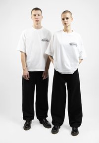 T-shirt bianchi oversize con logo nero, abbinate a pantaloni neri larghi. Le scarpe nere completano l'abbigliamento, strutturato e casual nel design.