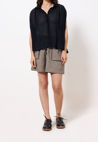 Femme portant un haut transparent noir à boutons, un short à carreaux beige et noir avec poches avant, et des chaussures à lacets noires avec semelles marron.