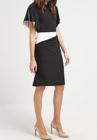 Svart klänning med fjärilskantade ärmar, vit midjebanddetalj, knälång nederkant och slät tygtextur. Matchad med nakna högklackade skor.