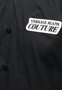 Vêtement noir avec deux boutons noirs et un écusson blanc affichant « VERSACE JEANS COUTURE » en texte noir en gras.