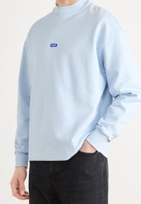 Felpa azzurra con collo alto, polsini a coste e un piccolo logo blu "HUGO" sul petto. Tessuto liscio, vestibilità comoda.