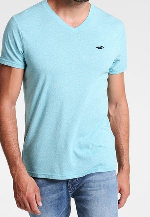 Hombre vistiendo una camiseta de manga corta con cuello en V de color azul claro con un pequeño logo de un pájaro negro en el pecho y jeans azules descoloridos.