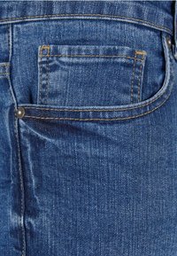 Gros plan sur un jean en denim bleu montrant la poche avant avec rivet et couture détaillée.