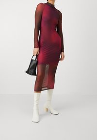 Rot schwarzes langärmliges Netzkleid mit figurbetonter Silhouette, hohem Kragen, gerafften Schultern und transparentem Material. Kombiniert mit einer schwarzen Tasche und weißen Blockabsatzstiefeln.