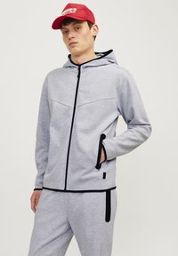 Svijetlosiva zip-hoodie s crnim detaljima, opremljena kapuljačom, bočnim džepovima i teksturiranom tkaninom. Uparena s odgovarajućim trenerkama.