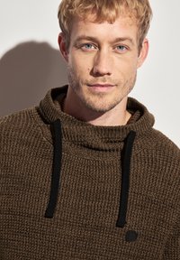 Hnědý pletený svetr s vysokým límcem a černými šňůrkami, který má texturovaný vzor a malé logo na přední straně.