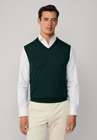 Ciemnozielony, bezrękawnik dzianinowy z dekoltem w serek, noszony na białej koszuli z kołnierzykiem, w zestawieniu z jasnymi spodniami.