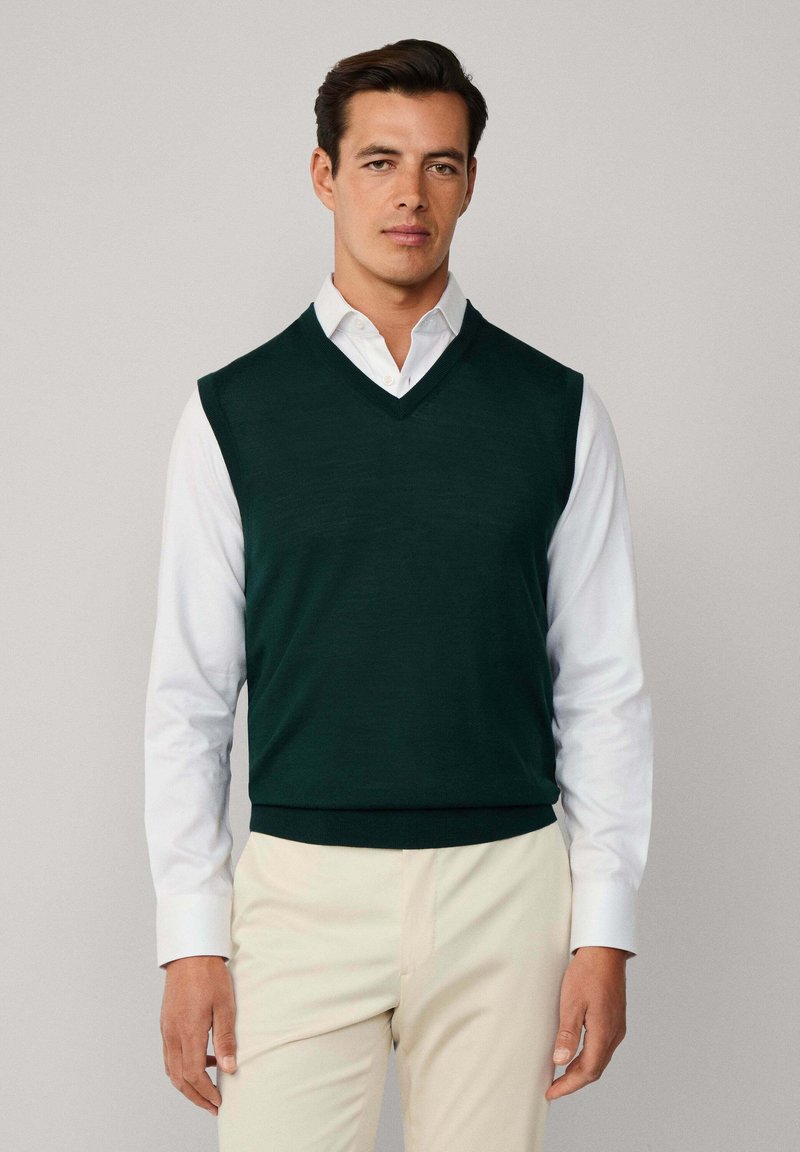 Ciemnozielony, bezrękawnik dzianinowy z dekoltem w serek, noszony na białej koszuli z kołnierzykiem, w zestawieniu z jasnymi spodniami.