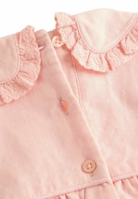 Vaaleanpunainen mekko, jossa on röyhelökaulus, nappikiinnitys ja hienovarainen ommeltu koriste. Puuvillakangas, jossa on tekstuuria.