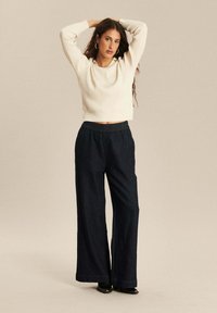 Crèmekleurige geribbelde trui en donkerdenim brede pijpen broek met een elastische tailleband, gecombineerd met zwarte hoge hakken, tegen een neutrale achtergrond.