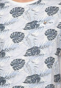 Chemise à manches courtes en tissu léger, avec un fond blanc et des imprimés de feuilles tropicales bleues et noires, ainsi qu'un petit détail de bouton marron.