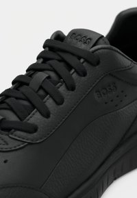 Zapatilla negra de cuero con parte superior texturizada, cordones planos, logo en relieve y detalles perforados en el costado. Diseño de perfil bajo.