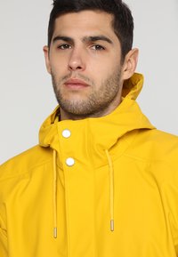 Gele waterdichte jas met hoge kraag, zilverkleurige hardware, elastische trekkoorden en een gladde, glanzende textuur.