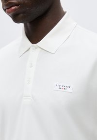 Κοντινή λήψη ενός ατόμου που φοράει λευκό πόλο με τρία κουμπιά και ένα σήμα Ted Baker Sport στο στήθος.