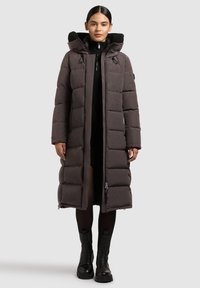 Cappotto lungo e imbottito di colore grigio scuro con zip frontale, cappuccio rimovibile e ampie sezioni trapuntate. Accentuato con pelliccia ecologica nera sul cappuccio.