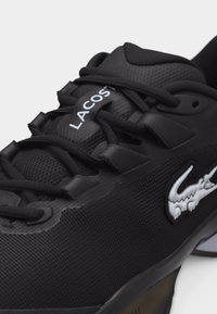 Μαύρο αθλητικό παπούτσι Lacoste με υφασμάτινη υφή, μαύρα κορδόνια, λευκό λογότυπο στο πλάι και "LACOSTE" στη γλώσσα του παπουτσιού.