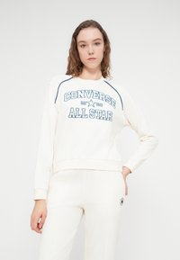 Sweat-shirt blanc avec texte graphique bleu "CONVERSE 1908 ALL STAR." Comprend un col rond et des poignets côtelés. Associé à un pantalon blanc assorti.