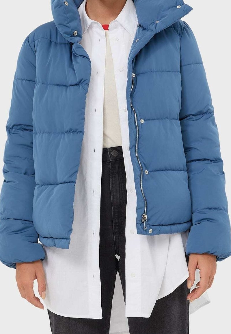 Person wearing a blue puffer jacket over a white button-down shirt, black pants, with hands relaxed at sides against a plain background.