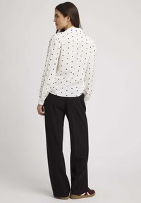 Chemise blanche à pois noirs, à manches longues, avec un col classique, associée à un pantalon noir à jambes larges et des chaussures à motifs marron et blanc.