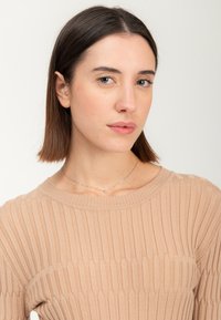 Junge Frau mit glattem, schulterlangem braunem Haar, die einen beigen, gerippten Pullover und eine filigrane Silberkette trägt, neutraler Hintergrund.