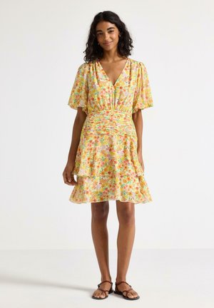 Femme portant une robe jaune fleurie à volants avec un col en V, des manches flottantes et des sandales plates marron, debout sur un fond blanc.
