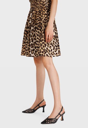 Donna che indossa un abito a più strati con stampa leopardata e décolleté slingback nere con dettagli cut-out su uno sfondo neutro.