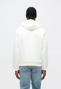 Sudadera con capucha blanca de corte holgado y capucha grande, hecha de tela suave. Combinada con vaqueros de mezclilla azul claro. Vista desde atrás.