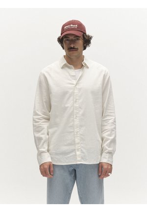 Muž so fúzmi, ktorý nosí červenú baseballovú čiapku, bielu košeľu na gombíky a svetlomodré džínsy na jednofarebnom bielom pozadí.