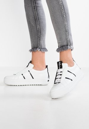 Persona que lleva zapatillas blancas de plataforma con rayas negras y vaqueros grises con dobladillo deshilachado, de pie sobre un suelo blanco y con fondo blanco.
