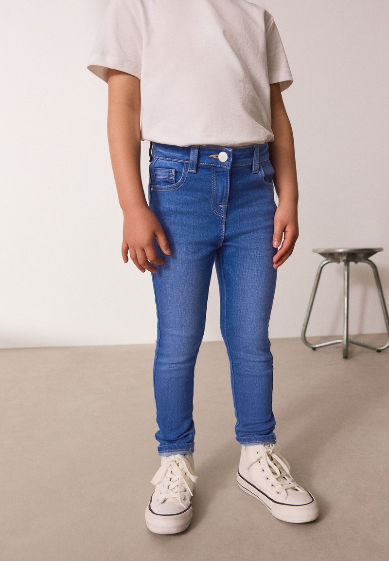 Jeans skinny in denim blu con tasche frontali, chiusura a bottone e cuciture a contrasto, abbinati a sneaker bianche e una t-shirt chiara.